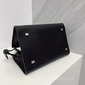 May include: A black leather handbag with a rectangular shape and a flap closure. The bag features silver-toned hardware on the corners and a thin strap. The bag is sitting on a white surface.