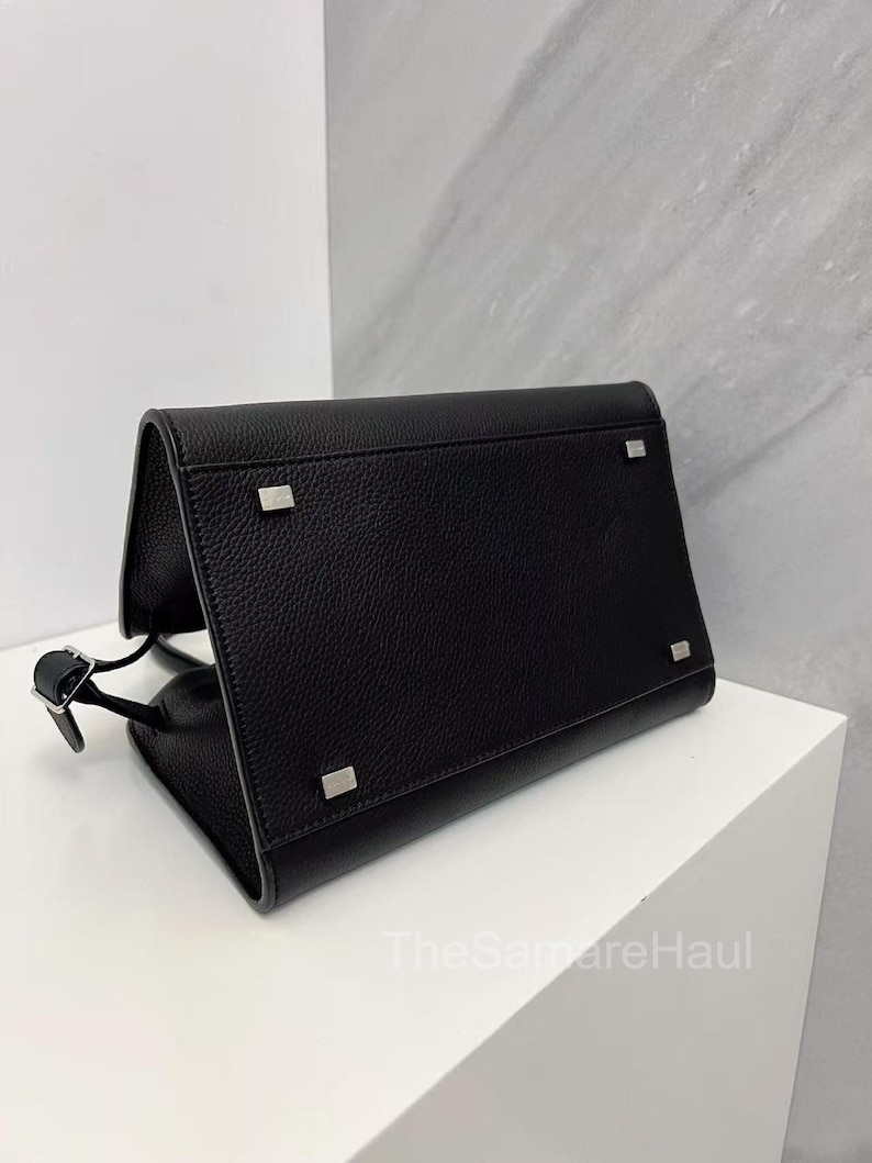 May include: A black leather handbag with a rectangular shape and a flap closure. The bag features silver-toned hardware on the corners and a thin strap. The bag is sitting on a white surface.