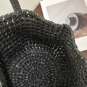 May include: A black woven tote bag with a braided handle. The bag has a textured, open weave design. The bag is made of a dark material, and the interior is visible through the weave.