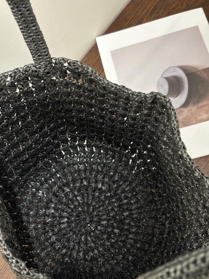 May include: A black woven tote bag with a braided handle. The bag has a textured, open weave design. The bag is made of a dark material, and the interior is visible through the weave.