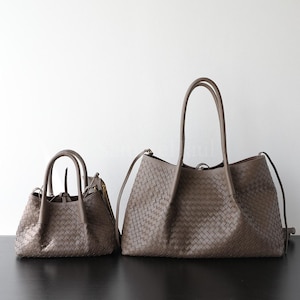 Puede incluir: Dos bolsos de mano de cuero tejido en un tono marr&oacute;n apagado. El bolso m&aacute;s grande tiene asas largas y una bandolera, mientras que el bolso m&aacute;s peque&ntilde;o tiene asas m&aacute;s cortas. Ambos bolsos est&aacute;n sobre un fondo blanco y una superficie negra.