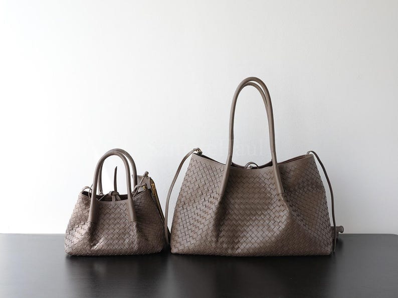 Puede incluir: Dos bolsos de mano de cuero tejido en un tono marr&oacute;n apagado. El bolso m&aacute;s grande tiene asas largas y una bandolera, mientras que el bolso m&aacute;s peque&ntilde;o tiene asas m&aacute;s cortas. Ambos bolsos est&aacute;n sobre un fondo blanco y una superficie negra.