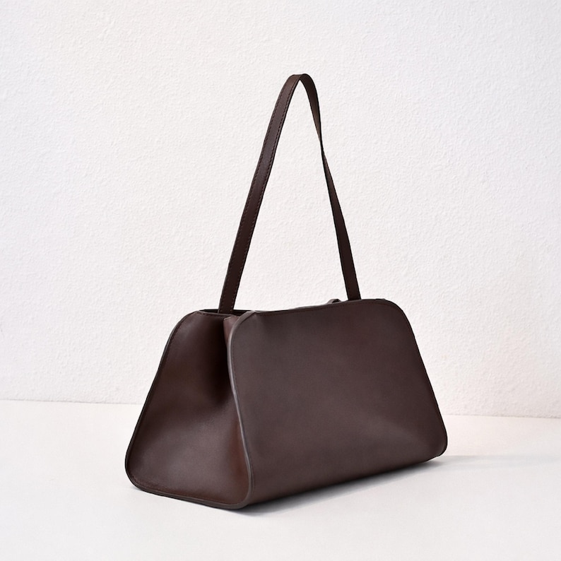 May include: A dark brown handbag with a rectangular body and a trapezoidal base. The bag has a single, long strap and is set against a white background. The bag's design is minimalist and sleek.
