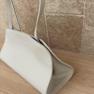 May include: A light gray handbag with a curved design and a shoulder strap. The bag is made of a smooth material and has a minimalist aesthetic. The bag is shown on a wooden surface.