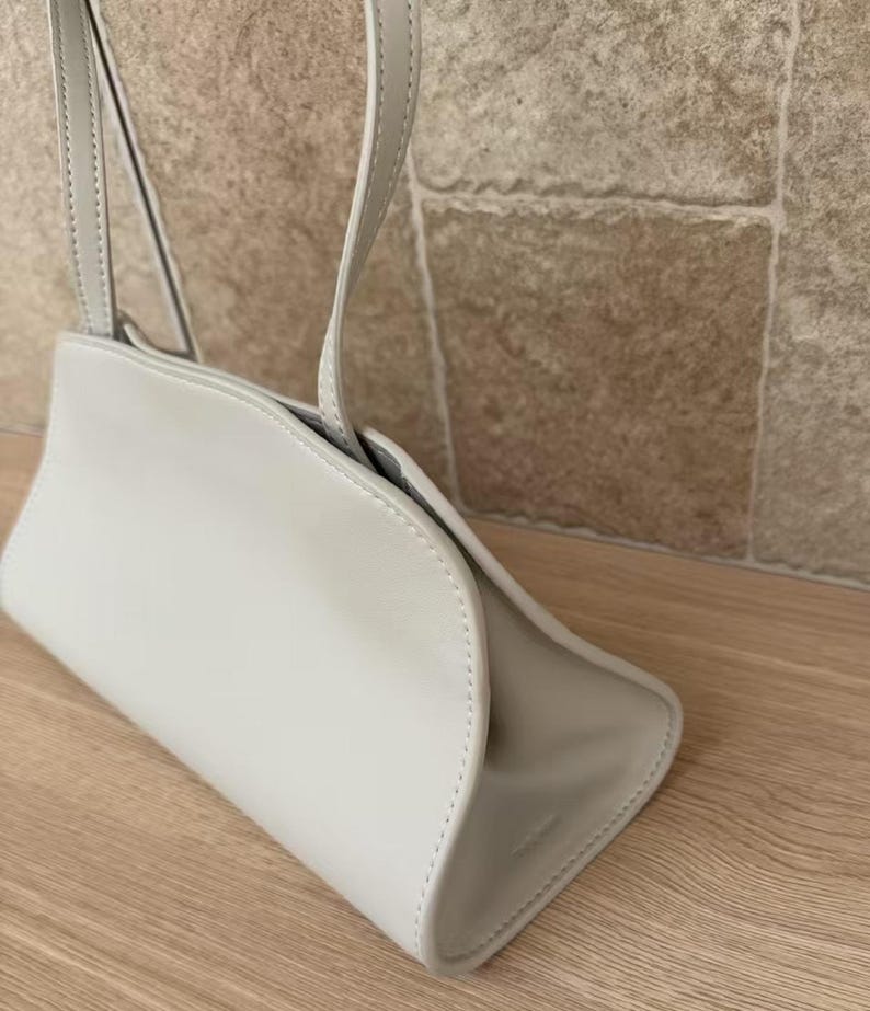 May include: A light gray handbag with a curved design and a shoulder strap. The bag is made of a smooth material and has a minimalist aesthetic. The bag is shown on a wooden surface.