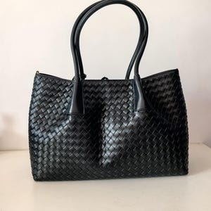 Puede incluir: Bolso tote de cuero negro tejido con dos asas curvas. El bolso tiene forma rectangular y un patr&oacute;n tejido. Las asas est&aacute;n unidas a la parte superior del bolso con detalles de cuero. El bolso est&aacute; sobre una superficie blanca.