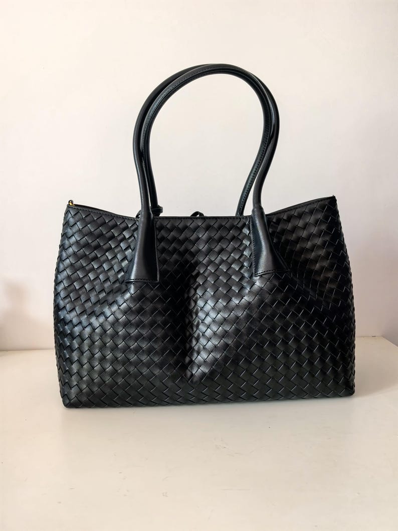 Puede incluir: Bolso tote de cuero negro tejido con dos asas curvas. El bolso tiene forma rectangular y un patr&oacute;n tejido. Las asas est&aacute;n unidas a la parte superior del bolso con detalles de cuero. El bolso est&aacute; sobre una superficie blanca.