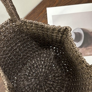 May include: A brown and tan woven tote bag. The bag features a textured, open weave design with a circular base and a braided handle. The interior of the bag is visible, showing the woven construction. The bag is suitable for carrying items.