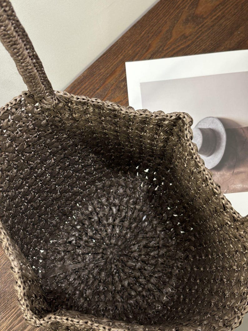 May include: A brown and tan woven tote bag. The bag features a textured, open weave design with a circular base and a braided handle. The interior of the bag is visible, showing the woven construction. The bag is suitable for carrying items.