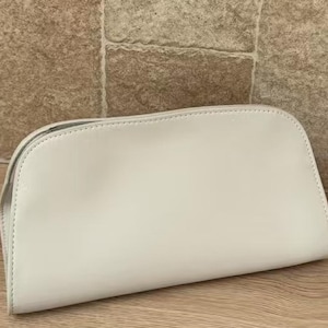 May include: A cream-colored, semi-circular cosmetic bag. The bag is made of a smooth material and has a zipper closure. The bag is sitting on a wooden surface with a stone background.