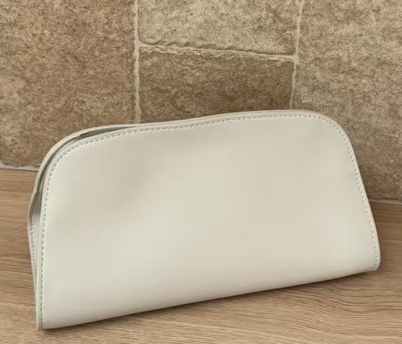 May include: A cream-colored, semi-circular cosmetic bag. The bag is made of a smooth material and has a zipper closure. The bag is sitting on a wooden surface with a stone background.