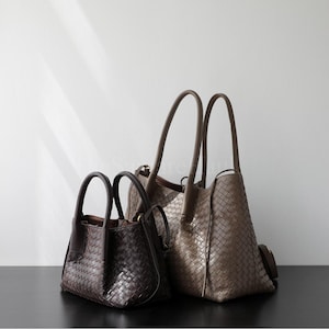 Puede incluir: Tres bolsos de mano de cuero tejido de diferentes tama&ntilde;os. El m&aacute;s peque&ntilde;o es marr&oacute;n oscuro, el mediano es marr&oacute;n claro y el m&aacute;s grande es de color topo. Todos tienen asas superiores y un patr&oacute;n tejido. Las bolsas se exhiben sobre una superficie oscura.