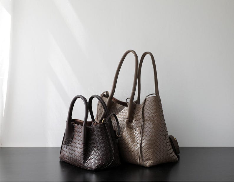 Puede incluir: Tres bolsos de mano de cuero tejido de diferentes tama&ntilde;os. El m&aacute;s peque&ntilde;o es marr&oacute;n oscuro, el mediano es marr&oacute;n claro y el m&aacute;s grande es de color topo. Todos tienen asas superiores y un patr&oacute;n tejido. Las bolsas se exhiben sobre una superficie oscura.