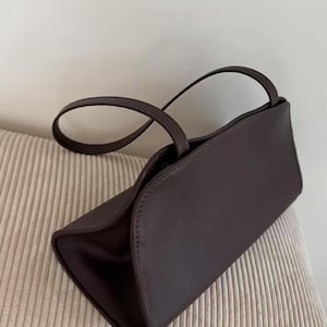 May include: A dark brown handbag with a rectangular shape and curved top. The bag has two thin handles and appears to be made of leather or a similar material. The bag is sitting on a beige, textured surface.