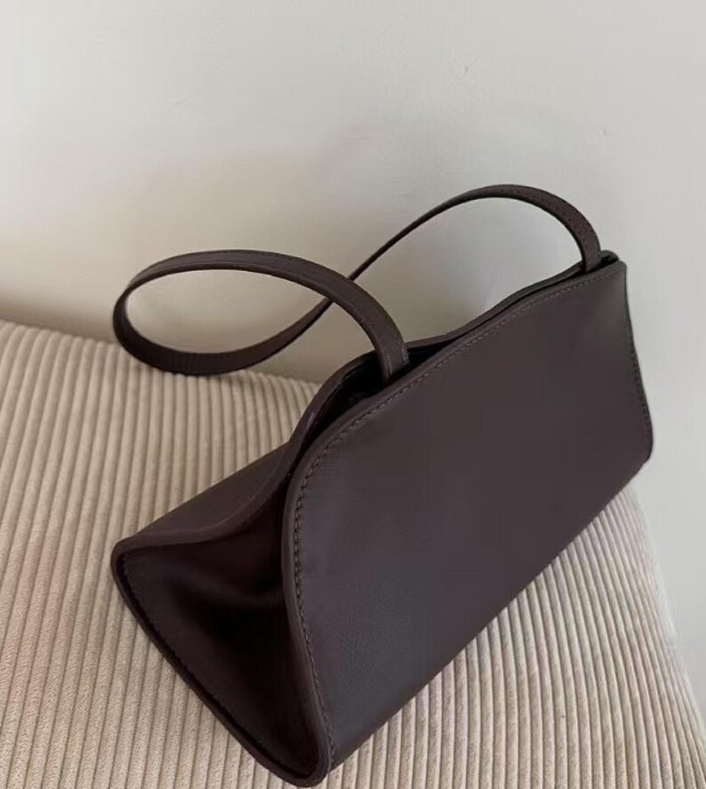 May include: A dark brown handbag with a rectangular shape and curved top. The bag has two thin handles and appears to be made of leather or a similar material. The bag is sitting on a beige, textured surface.