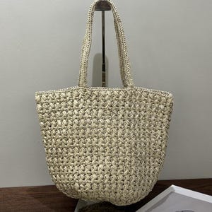 May include: A beige woven tote bag with two handles. The bag has a textured, basket-weave design. The handles are made of the same woven material. The bag is sitting on a stand.