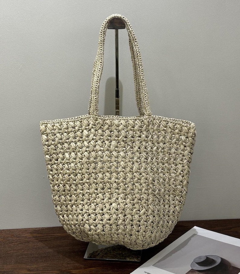 May include: A beige woven tote bag with two handles. The bag has a textured, basket-weave design. The handles are made of the same woven material. The bag is sitting on a stand.
