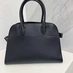 Può includere: Una borsa nera dal design strutturato. La borsa presenta due manici superiori arrotondati e una forma rettangolare con una parte superiore leggermente curva. Il materiale sembra avere una finitura strutturata. La borsa è su una superficie bianca.