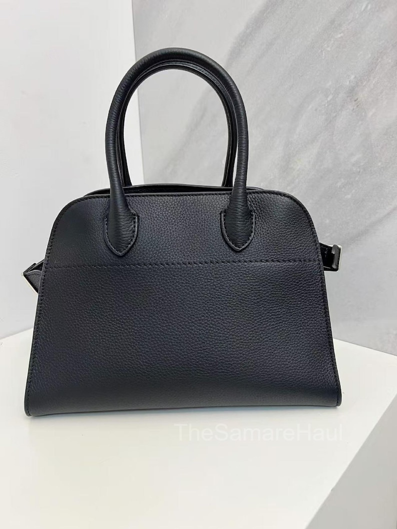 May include: A black handbag with a structured design. The bag features two rounded top handles and a rectangular shape with a slightly curved top. The material appears to have a textured finish. The bag is sitting on a white surface.