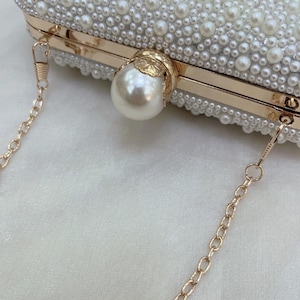 May include: A white beaded clutch bag with a gold chain strap. The rectangular bag is covered in small, white pearls and features a large pearl clasp. The gold frame adds elegance to this formal accessory, perfect for special occasions.