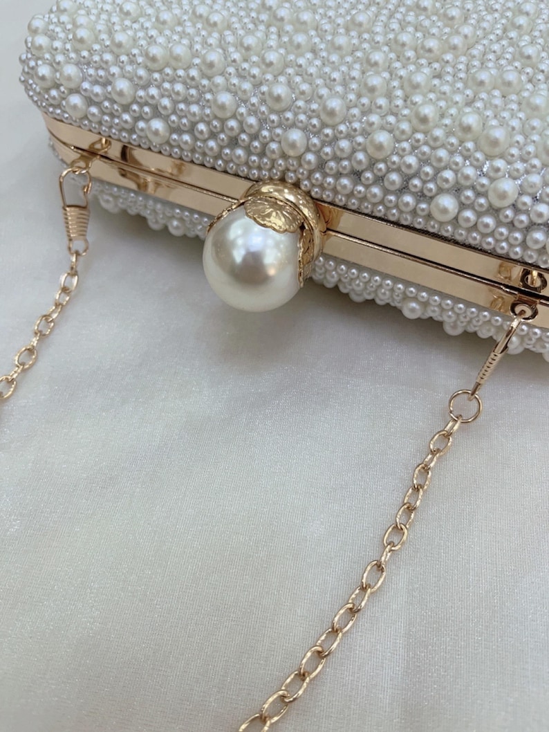 May include: A white beaded clutch bag with a gold chain strap. The rectangular bag is covered in small, white pearls and features a large pearl clasp. The gold frame adds elegance to this formal accessory, perfect for special occasions.