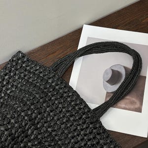 May include: Black woven tote bag with braided handles. The bag has a textured, open weave design. The handles are thick and sturdy. The bag is on a wooden surface next to a white paper with a hat illustration.
