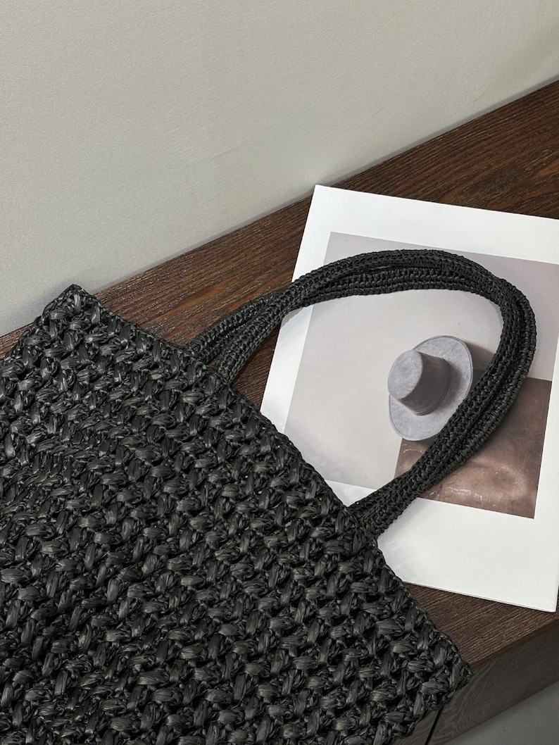 May include: Black woven tote bag with braided handles. The bag has a textured, open weave design. The handles are thick and sturdy. The bag is on a wooden surface next to a white paper with a hat illustration.