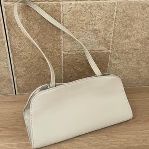 May include: A light-colored, rectangular handbag with a curved top and a thin shoulder strap. The bag is made of a smooth material and has a simple, minimalist design. The strap is attached to the bag with small metal rings.