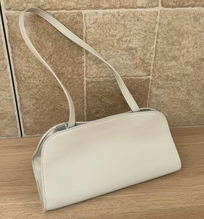 May include: A light-colored, rectangular handbag with a curved top and a thin shoulder strap. The bag is made of a smooth material and has a simple, minimalist design. The strap is attached to the bag with small metal rings.