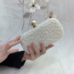 May include: A rectangular white clutch bag covered in small, round pearls. The bag has a gold-coloured clasp with a pearl accent. The background features floral decorations.