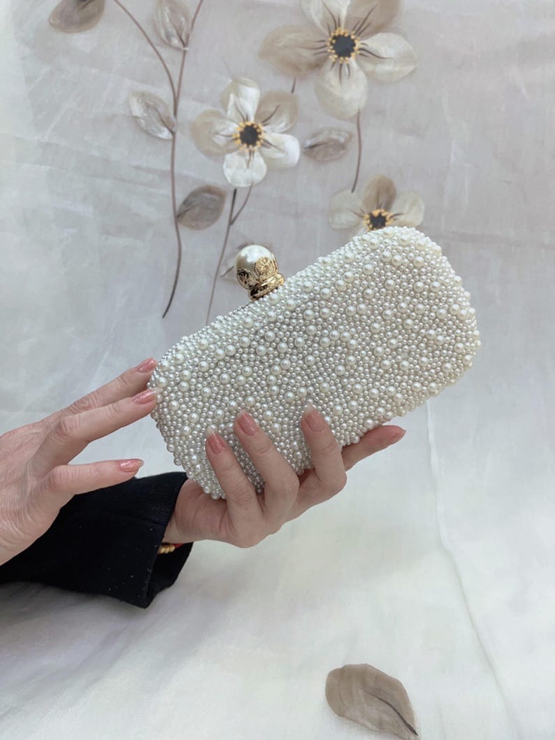 May include: A rectangular white clutch bag covered in small, round pearls. The bag has a gold-coloured clasp with a pearl accent. The background features floral decorations.