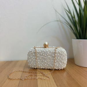 May include: A rectangular white beaded clutch bag with a gold chain strap. The bag has a gold clasp and is covered in small, round, white beads. The gold chain is draped across a wooden surface.