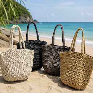 May include: Four woven tote bags in beige, black, and brown hues are displayed on a sandy beach. Each bag has a matching handle and a spacious interior. The bags are ideal for carrying beach essentials.