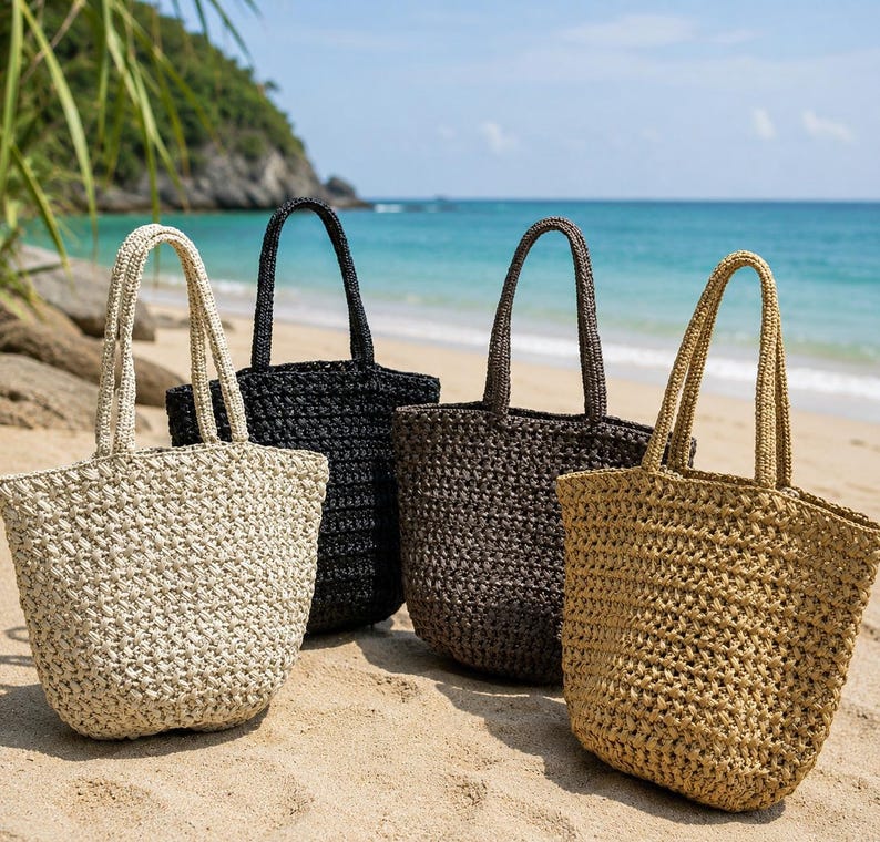 May include: Four woven tote bags in beige, black, and brown hues are displayed on a sandy beach. Each bag has a matching handle and a spacious interior. The bags are ideal for carrying beach essentials.