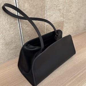 May include: A black handbag with a rectangular body and curved edges. The bag has two handles and a flap closure. The bag is made of a smooth material and sits on a wooden surface.