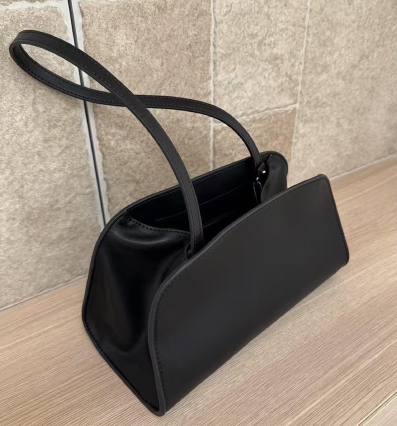 May include: A black handbag with a rectangular body and curved edges. The bag has two handles and a flap closure. The bag is made of a smooth material and sits on a wooden surface.