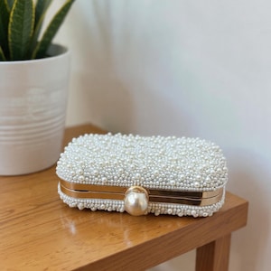 May include: A rectangular white clutch bag, covered in small, round pearls. It features gold-coloured metal trim and a large pearl clasp. The bag is on a wooden surface.