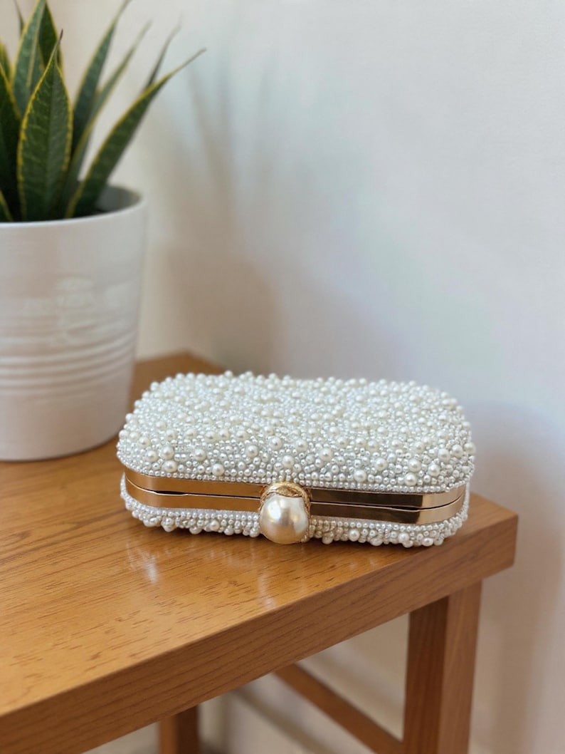 May include: A rectangular white clutch bag, covered in small, round pearls. It features gold-coloured metal trim and a large pearl clasp. The bag is on a wooden surface.
