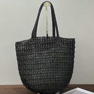 May include: A black woven tote bag with two handles. The bag has a rounded base and a textured surface. The handles are made of the same woven material as the bag. The bag is displayed on a wooden surface.