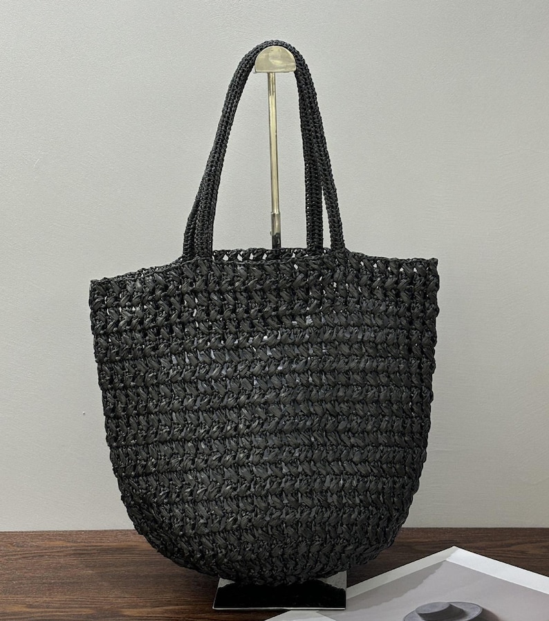 May include: A black woven tote bag with two handles. The bag has a rounded base and a textured surface. The handles are made of the same woven material as the bag. The bag is displayed on a wooden surface.