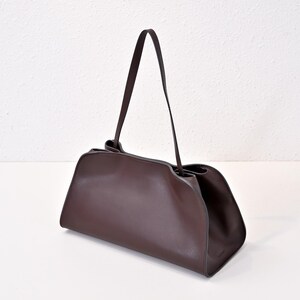 May include: A dark brown leather handbag with a rectangular base and a curved top. The bag has a long, thin strap and a minimalist design. The bag is made of smooth leather and has a simple, elegant look.