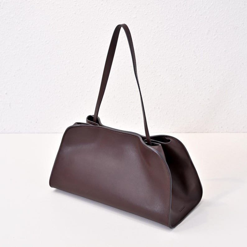 May include: A dark brown leather handbag with a rectangular base and a curved top. The bag has a long, thin strap and a minimalist design. The bag is made of smooth leather and has a simple, elegant look.