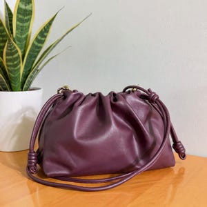 May include: A plum-colored leather handbag with a gathered design. The bag has a drawstring closure and a knotted handle. The bag is on a wooden surface, with a potted plant in the background.