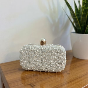 May include: A rectangular, off-white clutch covered in small, round, faux pearls. The clasp is a gold-coloured sphere. The clutch is sitting on a wooden surface.