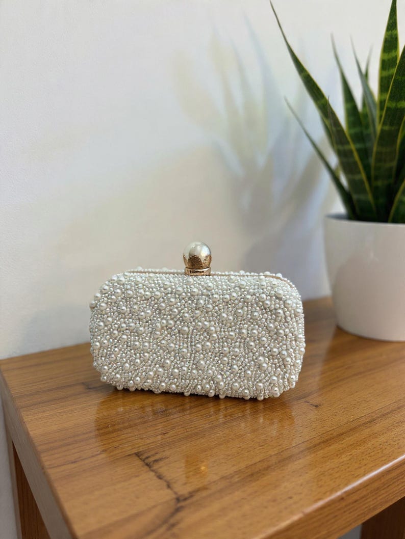 May include: A rectangular, off-white clutch covered in small, round, faux pearls. The clasp is a gold-coloured sphere. The clutch is sitting on a wooden surface.