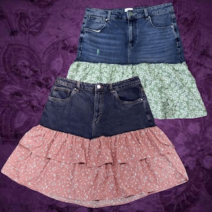 May include: Two denim skirts with floral ruffle hems. One skirt has a light wash denim top with a green floral ruffle. The other skirt has a dark wash denim top with a pink floral ruffle.