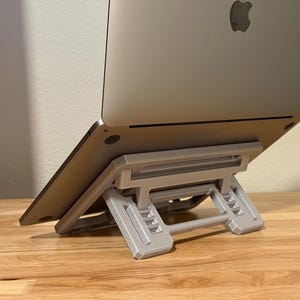 May include: A silver laptop computer is propped up on a gray laptop stand. The stand is made of plastic and has a geometric design. The laptop is angled upward, displaying the Apple logo on the back. The stand is on a wooden surface.