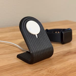 May include: A black, 3D-printed Apple Watch charging stand with a white charging puck. The stand has a curved design with vertical ridges. A black Apple Watch with a black strap rests nearby. A white charging cable is connected.