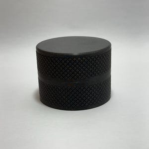 Op de afbeelding: Een cilindrisch, donkergrijs object met een getextureerd, ruitvormig oppervlak. De bovenkant is plat en glad. Het object lijkt van metaal te zijn en is waarschijnlijk een onderdeel of deel van een groter item.