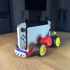 May include: A white and black handheld gaming console docked in a red, white, and blue stand shaped like a race car. The stand features four black tyres with yellow rims and a red cylindrical component on the side with the word FUEL embossed on it.
